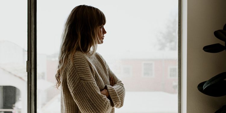 Woman looking sadly out of a window with her arms crossed.