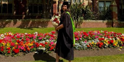 Alexandra in a black cap and gown stands in front of a vibrant flower bed. She is holding a large bouquet of flowers and is looking towards the camera with a slight smile. The background features a red brick building and various green plants.