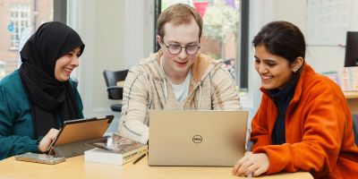 Three students sitting around a table looking at a laptop. They are smiling.