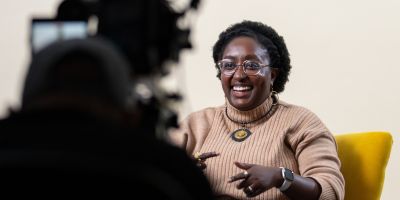 Damarie sits on a yellow chair in front of a white background, she is smiling to a video camera.