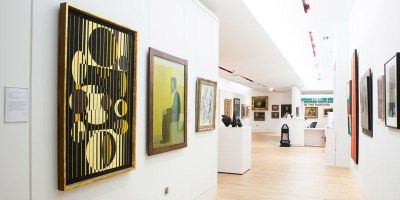 Decorative image - photograph inside Stanley and Audrey Burton Gallery at Leeds. Viewpoint: looking down long narrow gallery room, large paintings hung on white walls in well-lit room with light wood floors.