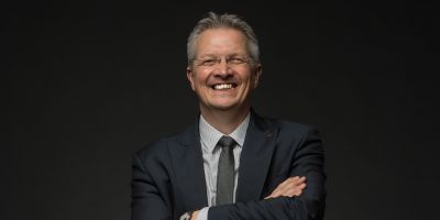 Nick Taylor stands against a dark background, he has his arms crossed and he is smiling.