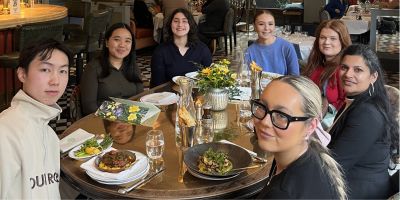 A group of seven people sitting around a round table in a restaurant, smiling at the camera. The table has food, drinks, and a floral centerpiece.