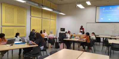 A lecturer standing behind a desk at the front of a large classroom facing 11 students sitting in small clusters.