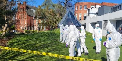People in forensic suits investigate a mock crime scene on a grassy lawn, surrounded by yellow tape, near modern and historic university buildings.