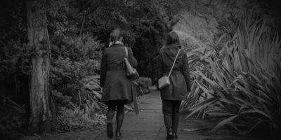 A greyscale photograph of two women walking in a park. We see them from behind.
