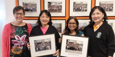 Four friends smiling at the camera while holding two graduation photos from 30 years ago