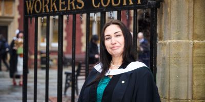 Sarah Lloyd stands in graduation gown in front of Clothworkers Court, redbrick buildings and decorated railings visible behind her.