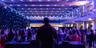 A DJ is stood facing forwards in front of a turntable, playing music to a large group of people who are dancing and talking. The room is lowly lit by spotlights.