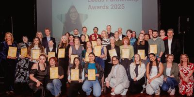 Group of award winners and attendees holding certificates at the Leeds Partnership Awards 2025, posed in front of a screen displaying the event title and a featured portrait.