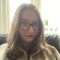 Rebecca is sat in a black leather armchair. She has long brown hair, blue eyes, and wears thick rimmed tortoise shell glasses. She is wearing a pink jumper, and a grey jacket