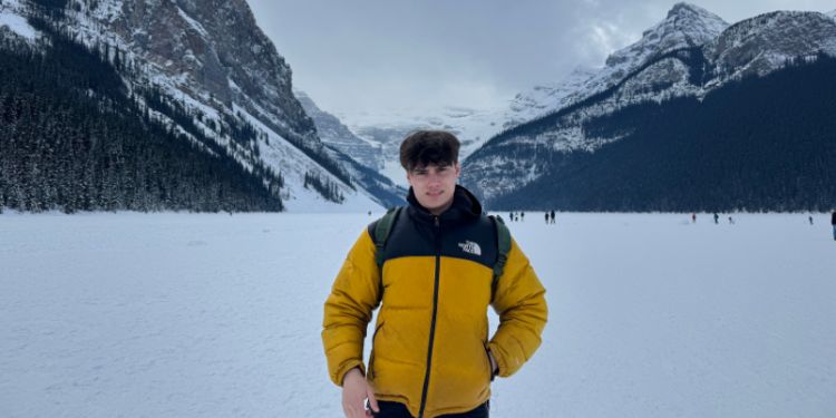 A young man stands on a snowy landscape with mountains in the background. He is wearing a yellow and black North Face jacket and has a backpack on. The sky is cloudy, and the scene is surrounded by snow-covered trees and mountains.