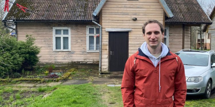 Matth is standing in front of an old wooden house with a gabled roof. The man is wearing a red jacket with a white hoodie underneath. The house has two small windows with white frames and a door in the centre. There is a green lawn in front of the house. 