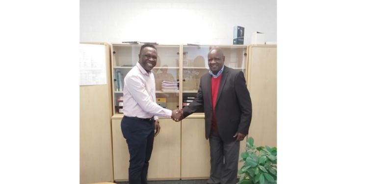 Two professors smiling and shaking hands, looking to camera, in a room stood in front of a bookcase.